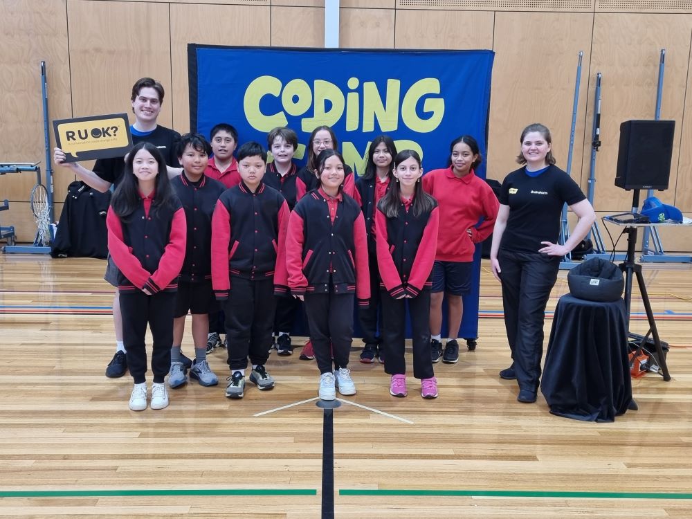 A photo taken during the week of R U OK?Day 2023. Two Brainstorm Productions actors are posing for a photo with a group of primary school students. One actor is wearing an R U OK? shirt and holding an R U OK? sign.