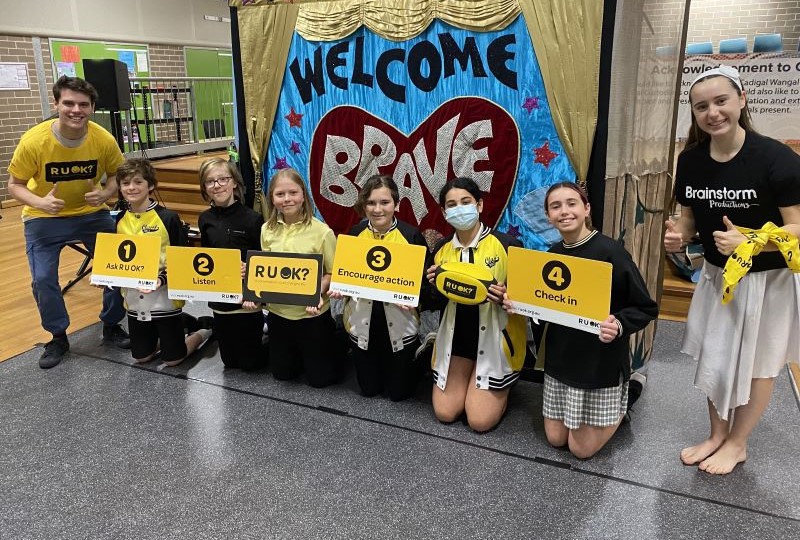 A photo taken during the week of R U OK?Day 2023. Two Brainstorm Productions actors are posing for a photo with a group of primary school students. One actor is wearing an R U OK? shirt and the students are holding R U OK? signs, with the 4 R U OK? steps.