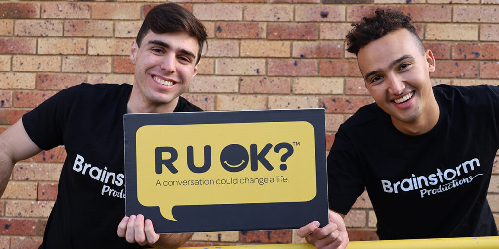 Two Brainstorm Productions actors are holding an R U OK? sign and leaning forward, smiling at the camera