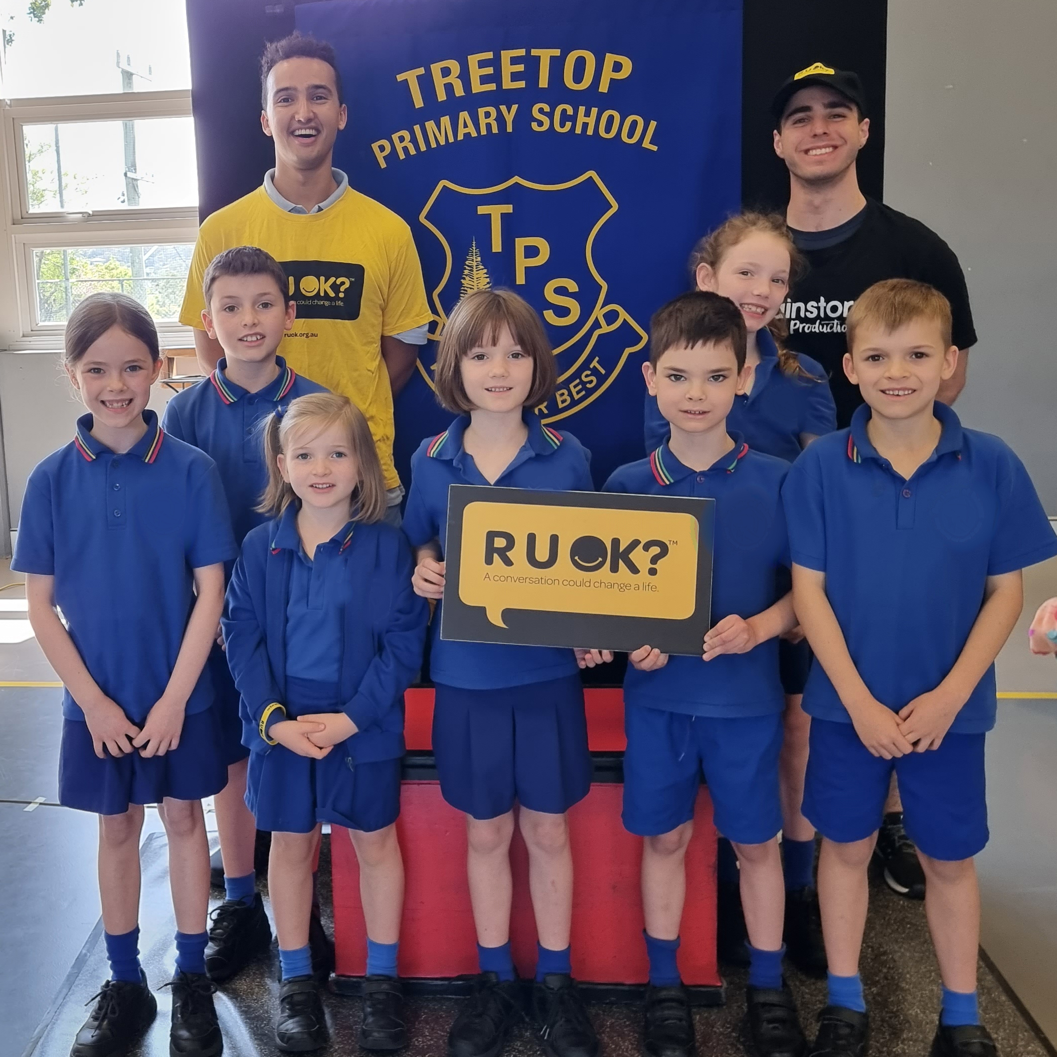 A photo taken during the week of R U OK?Day 2023. Two Brainstorm Productions actors are posing for a photo with a group of primary school students. One actor is wearing an R U OK? shirt and the students are holding an R U OK? sign.