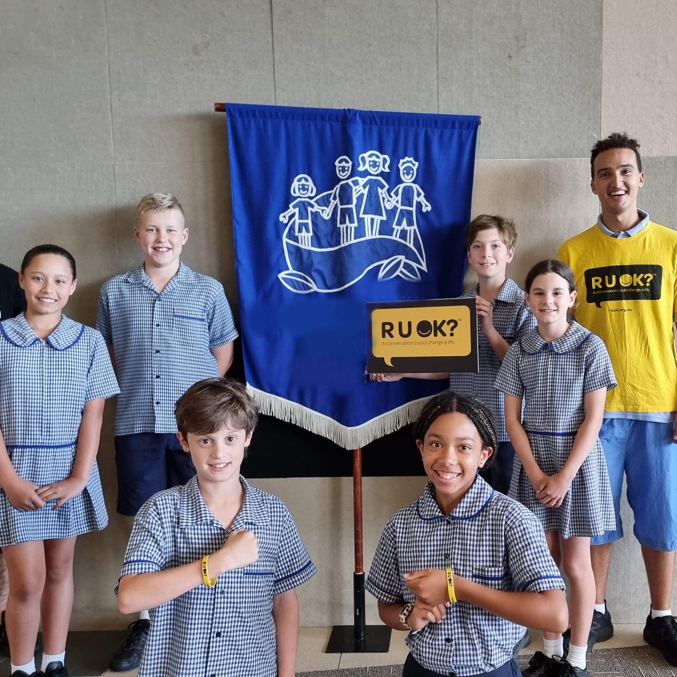 A photo taken during the week of R U OK?Day 2023. A Brainstorm Productions actor is posing for a photo with a group of primary school students. The actor is wearing an R U OK? shirt and the students are holding an R U OK? sign.