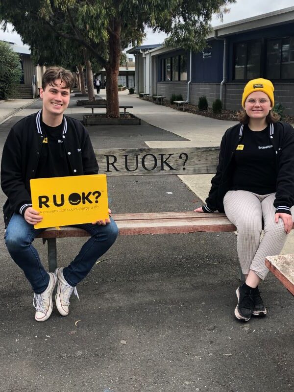 Two Brainstorm actors are sitting on a bench at Lalor Secondary College that says "R U OK?"