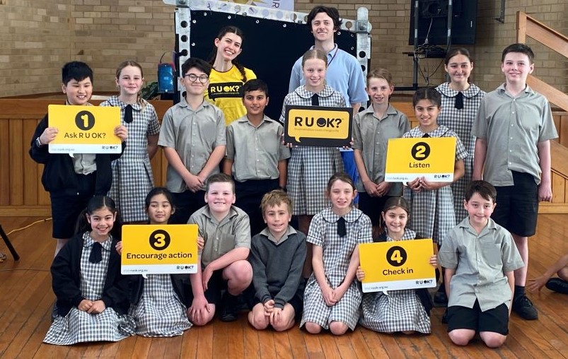 A photo taken during the week of R U OK?Day 2023. Two Brainstorm Productions actors are posing for a photo with a group of primary school students. One actor is wearing an R U OK? shirt and the students are holding R U OK? signs, with the 4 R U OK? steps.