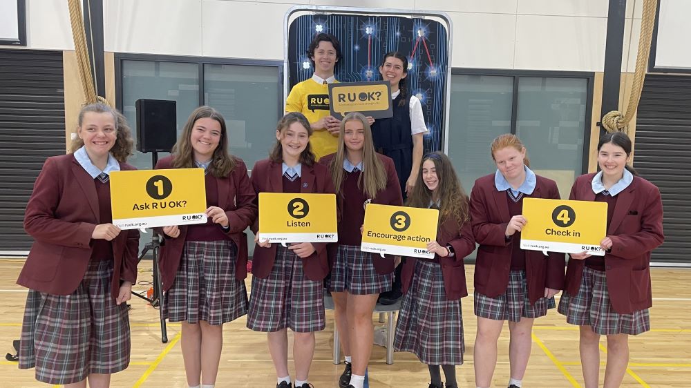 A photo taken during the week of R U OK?Day 2023. Two Brainstorm Productions actors are posing for a photo with a group of high school students. One actor is wearing an R U OK? shirt and the students are holding R U OK? signs, with the 4 R U OK? steps.