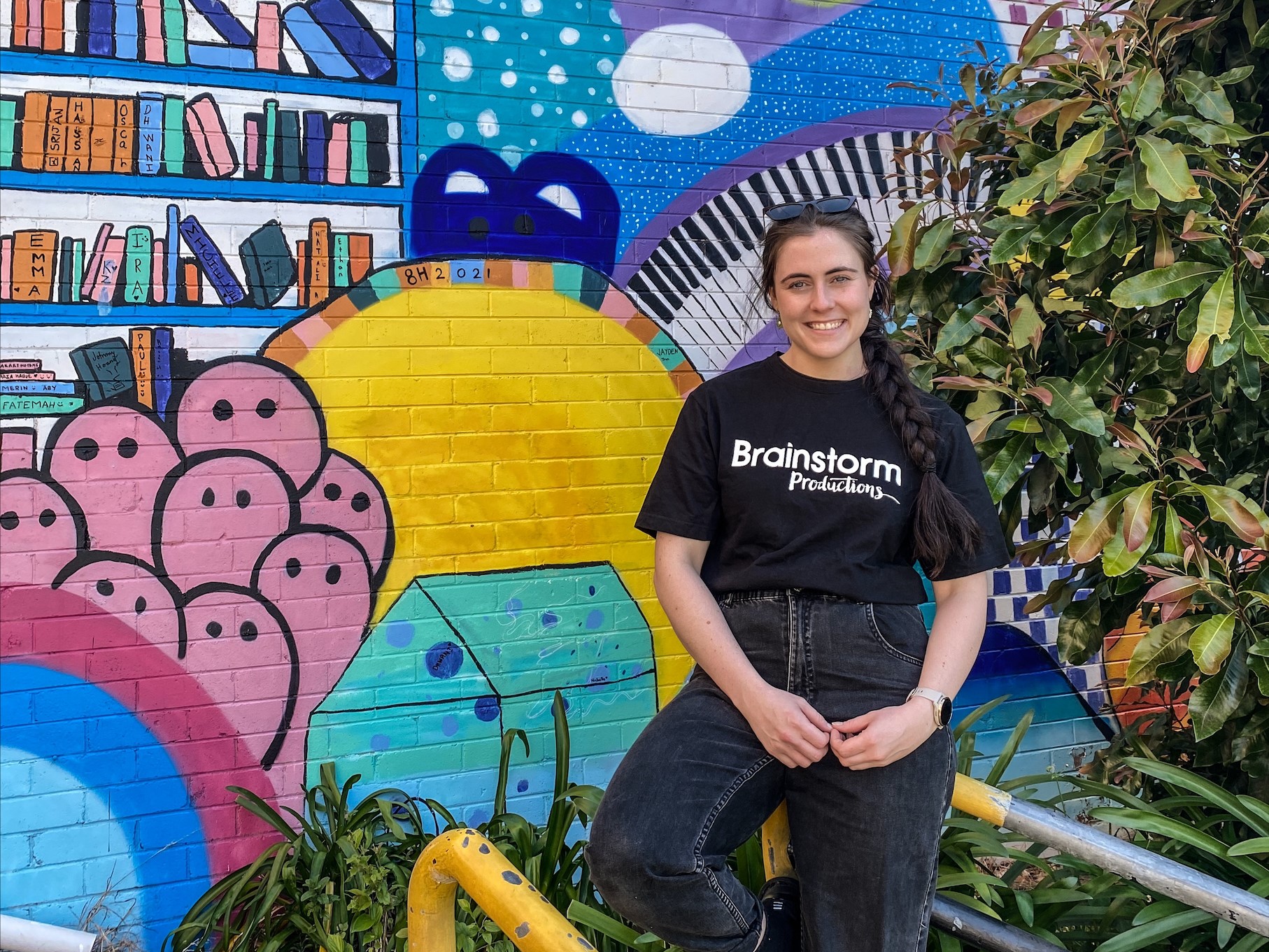A Brainstorm Productions actor is wearing a Brainstorm shirt and standing in front of a colourful mural at Kogarah High School, NSW.