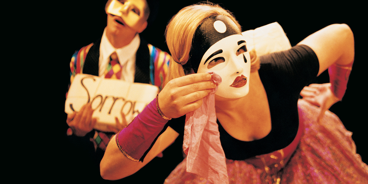 A still from an early version of the performance of the show 'Buddies' by Brainstorm Productions. Two actors are dressed in Commedia Dell'arte costume. They are both wearing masks and one is holding a block that says 'sorrow'.