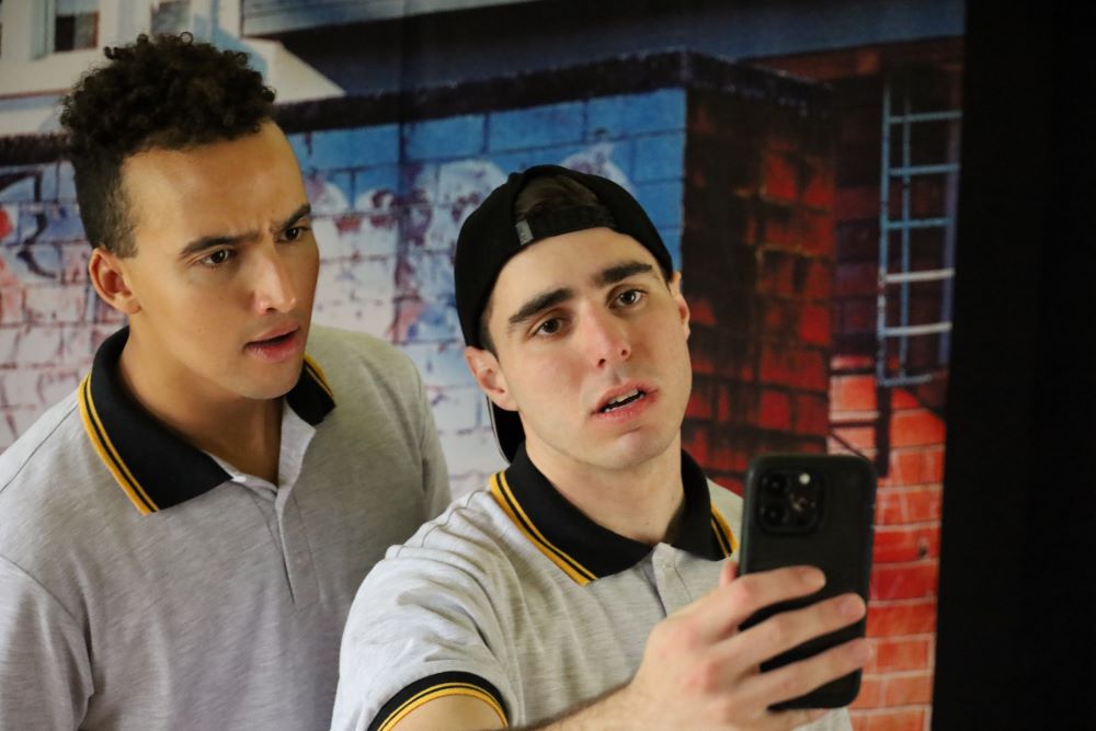A still from the high school performance 'Sticks & Stones Years 7-11' by Brainstorm Productions. One actor is taking a photo of someone outside the frame and the other actor is looking at the photo with concern.