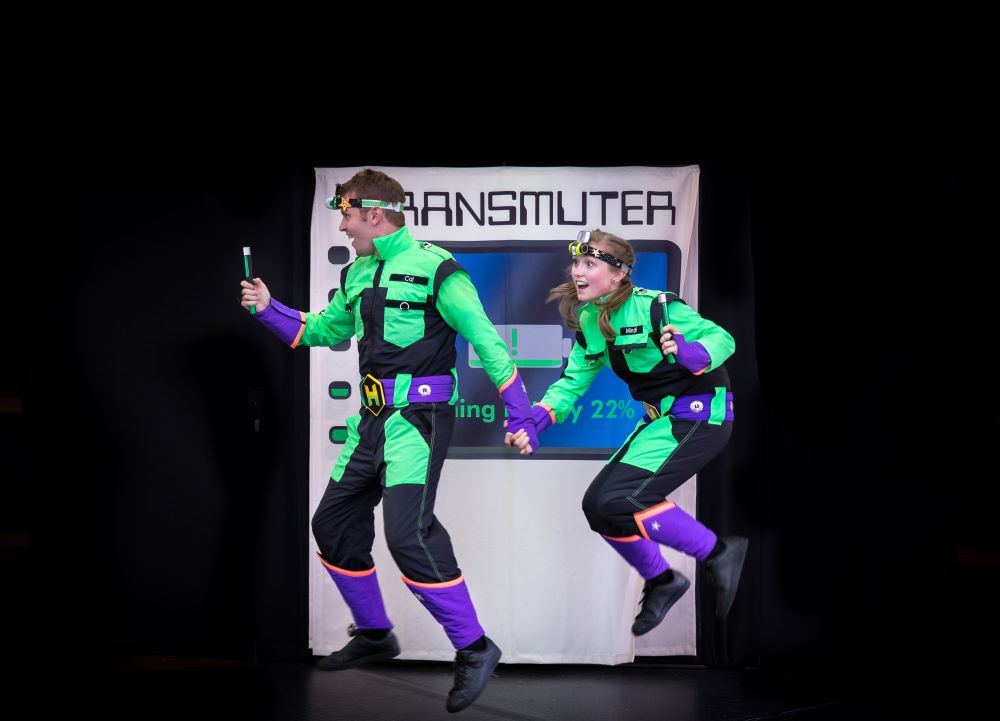 A still from the current, updated version of The H Team. Two actors are dressed in colourful jumpsuits, holding hands and jumping in the air in front of a backdrop that says 'Transmuter'.