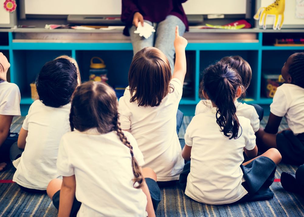 Young children are sitting on the floor in a classroom facing the teacher