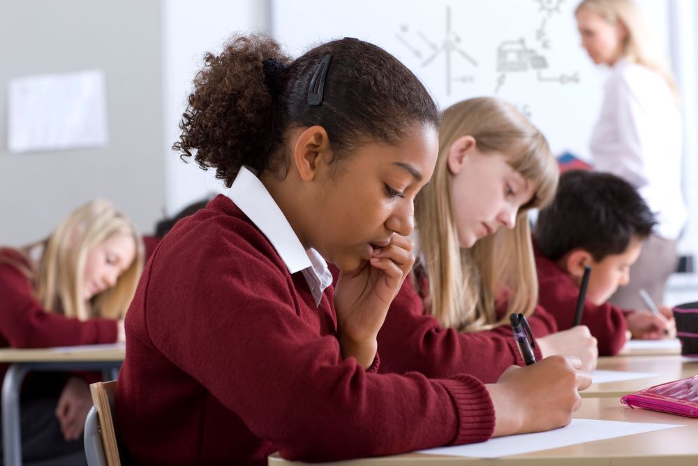 High school students are sitting at their desks writing and the student in the foreground is looking concerned.