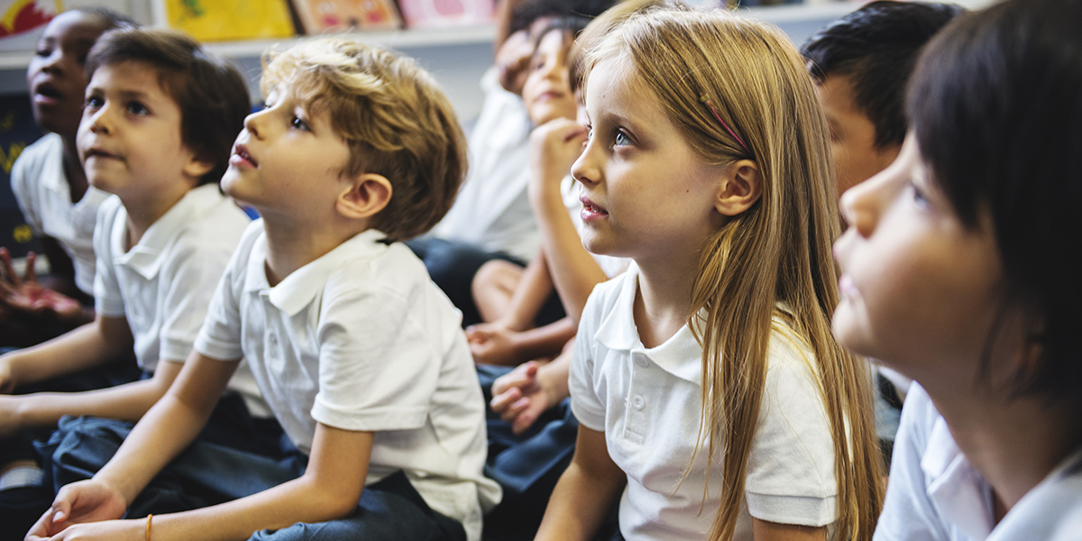 A group of primary school students are sitting on the classroom floor and listening to a teacher.