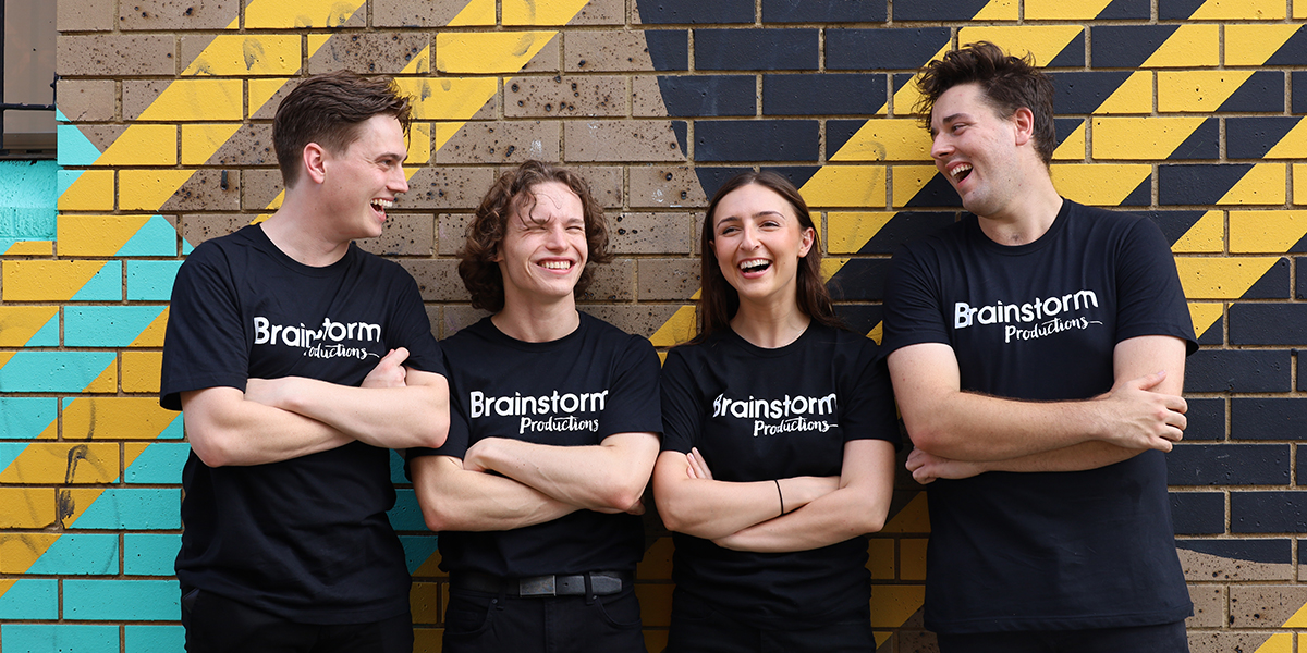 Four actors wearing Brainstorm productions t-shirts are standing together in front of a colourful mural with arms folded and laughing
