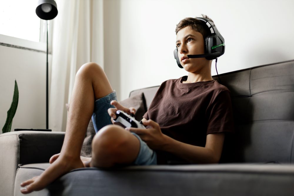 A young man is sitting on the couch holding a gaming controller and wearing a headset
