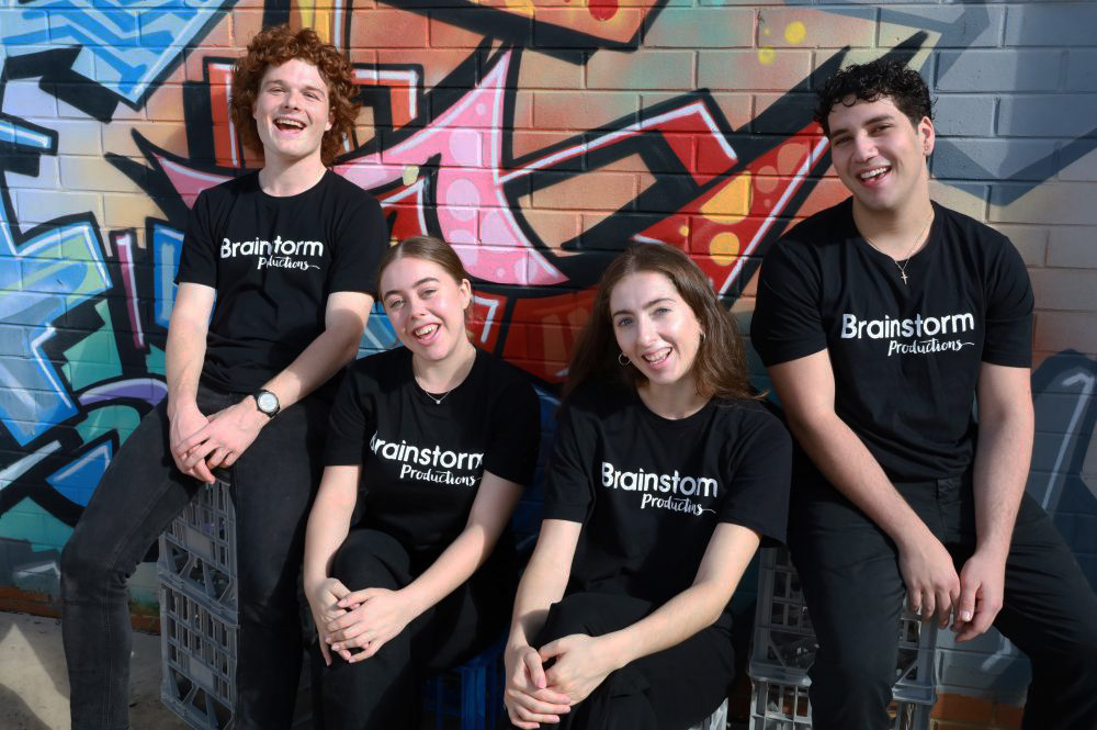 A promo image of four Brainstorm actors wearing company t-shirts and sitting in front of a colourful street art wall.
