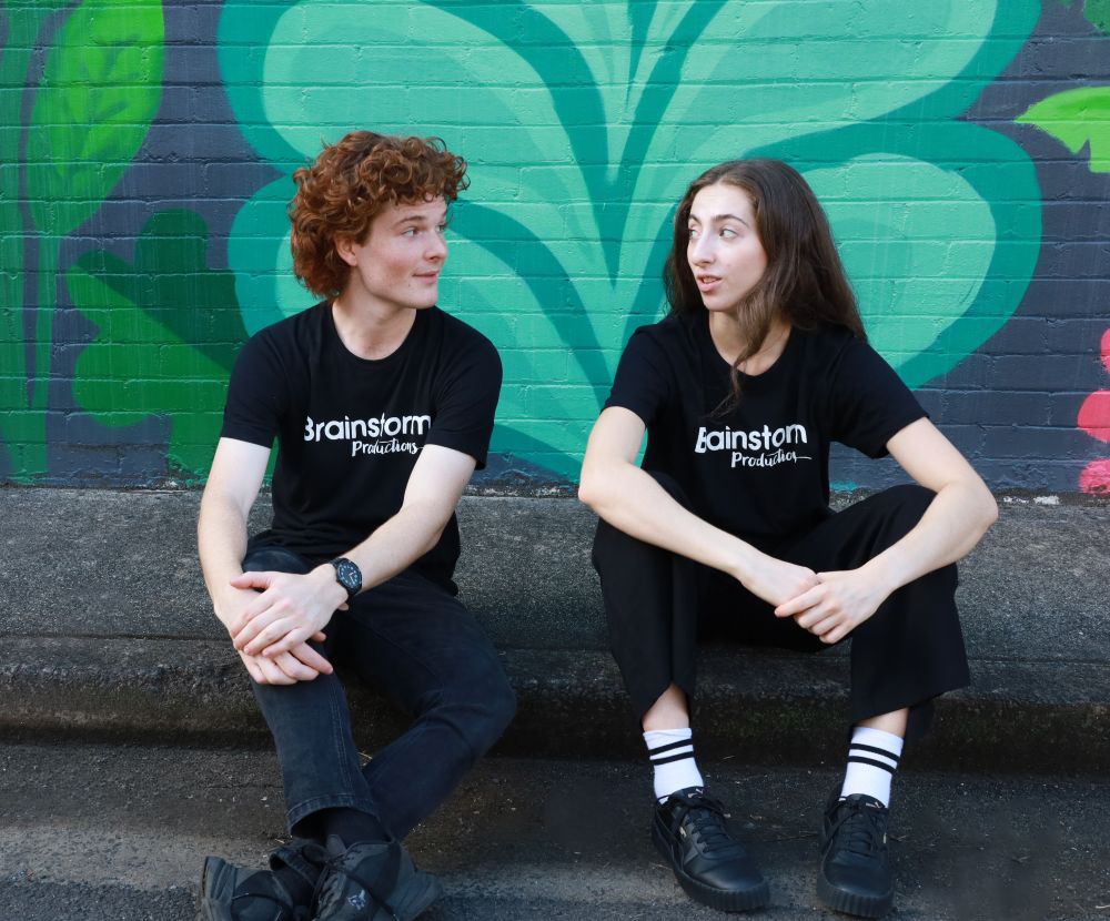 Two Brainstorm actors sit on the kerb in front of a brightly coloured street art mural, looking at each other and having a conversation