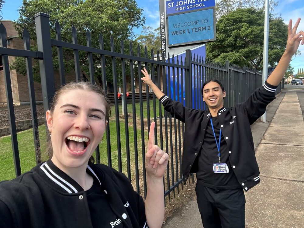 Two Brainstorm actors outside St Johns Park High School with big smiles, pointing to a sign that says 'Week 1, Term 2'