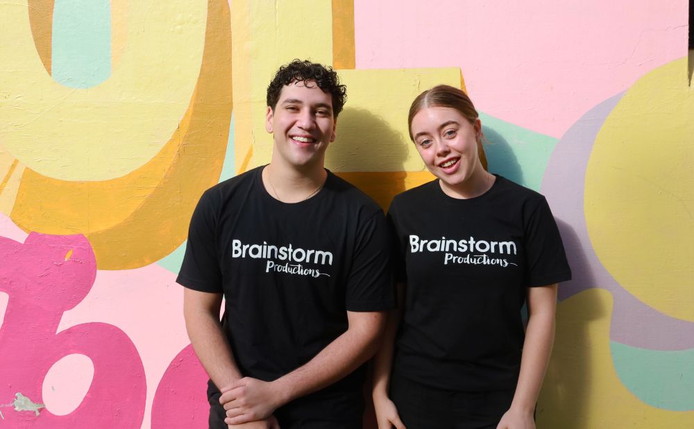 Two Brainstorm Productions actors are standing in front of a bright street art mural wearing Brainstorm t-shirts and smiling at the camera with friendly, approachable expressions