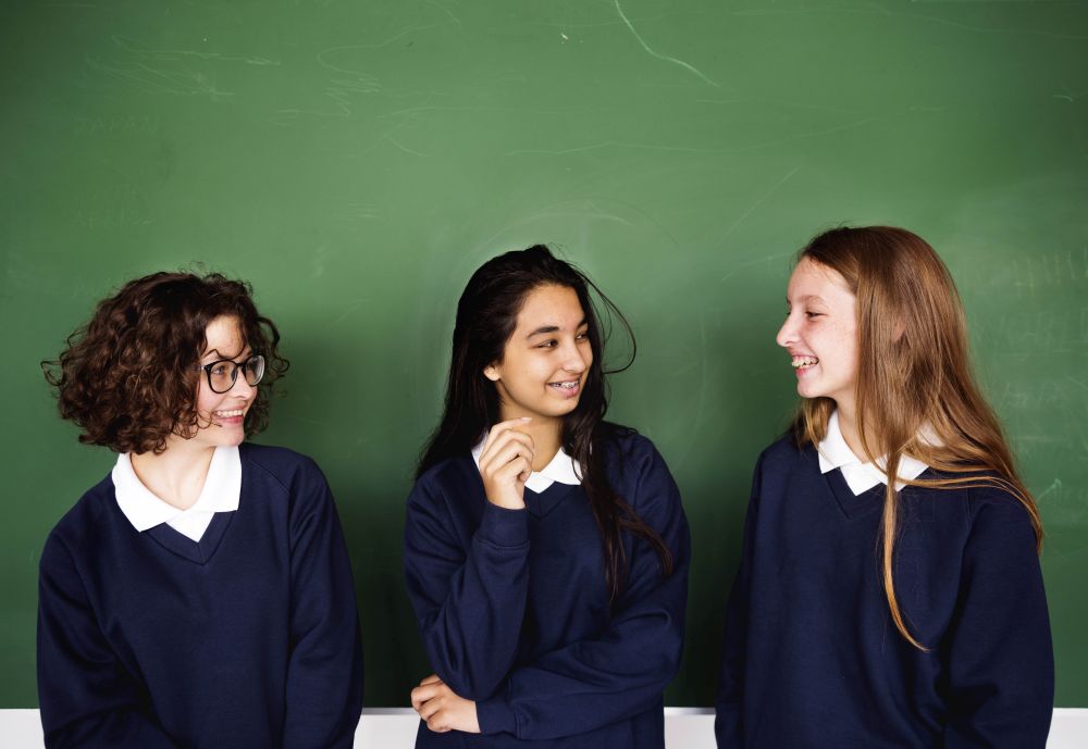 Three high school students are standing in front of a blackboard smiling at each other