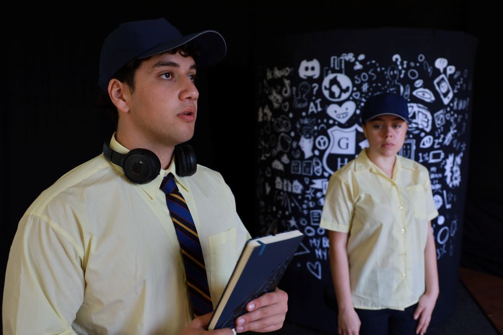 Two Brainstorm actors are in a moment from the high school performance Verbal Combat. One actor is in the foreground holding a book and looking at someone off camera. The other actors is standing in the background looking at the booking with a worried expression.