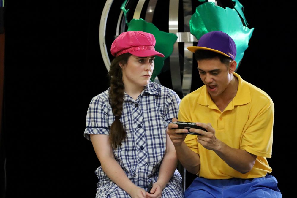 Two Brainstorm actors during a performance of 'The Human Race', a primary school show that addresses bullying, online safety, values and belonging. One actor is on a device looking angry and aggressive, the other actor is looking at him with a concerned expression.