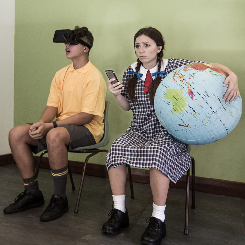 Promo image for primary school show The Human race - two young actors are sitting in chairs, one is holding a globe and a phone looking worried, the other is wearing a VR headset.