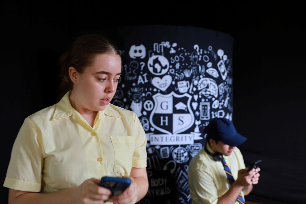 A still from the high school performance Verbal Combat, two actors are on on their phones, one in the foreground looks concerned.