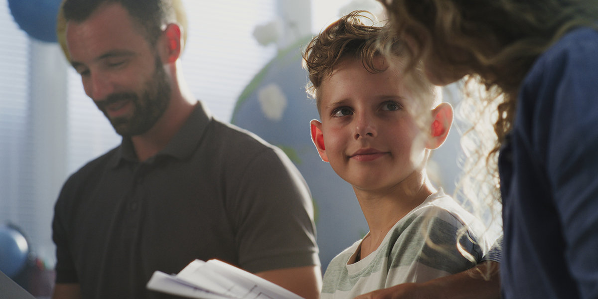 A young teenager is smiling at an adult, with another adult standing near them, illustrating the importance of support from trusted adults