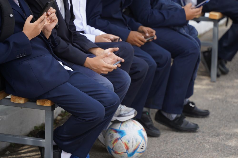 A group of high school aged boys sitting on a bench with many on their phones.