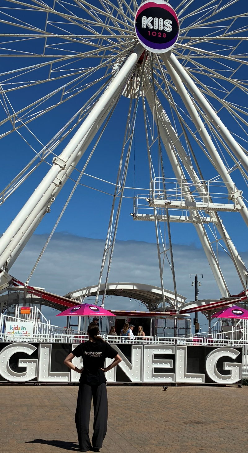 A Brainstorm productions actor stands in front of the ferris wheel at Glenelg Adelaide