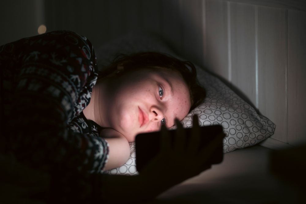 A young teenager is looking at her phone at night with an unhappy expression, showing the potential impact of AI and social media on mental health in vulnerable adolescents